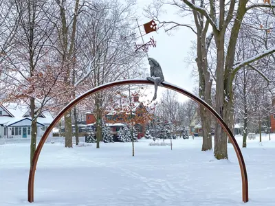 Sculpture of a young boy perched on a large arch holding a wind vane in a snowy park